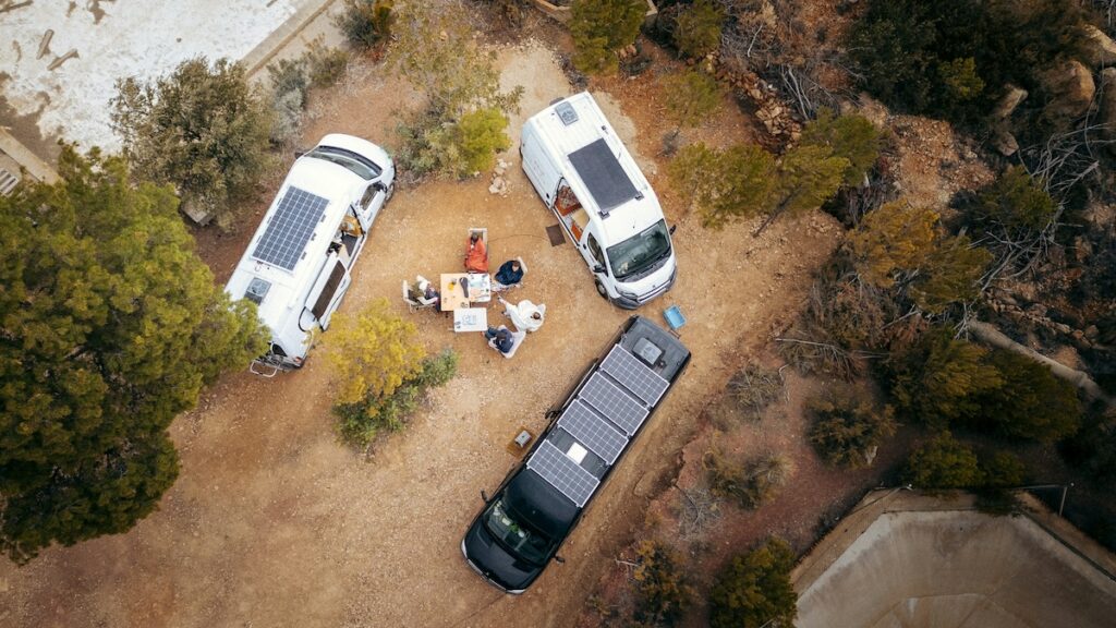 Aerial view of three camper vans with rooftop solar panels parked in a dirt clearing as people sit around a table outdoors.