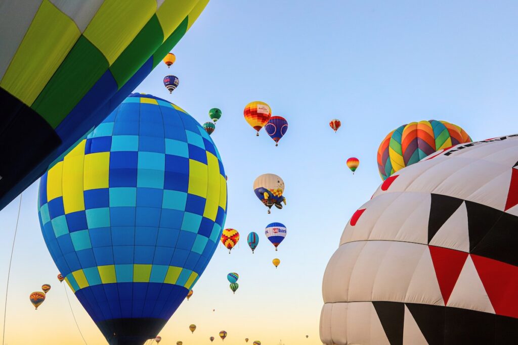 Multiple colorful hot air balloons inflated and floating in a clear blue sky, with a large blue-yellow checkered balloon foreground and another patterned balloon to the right.