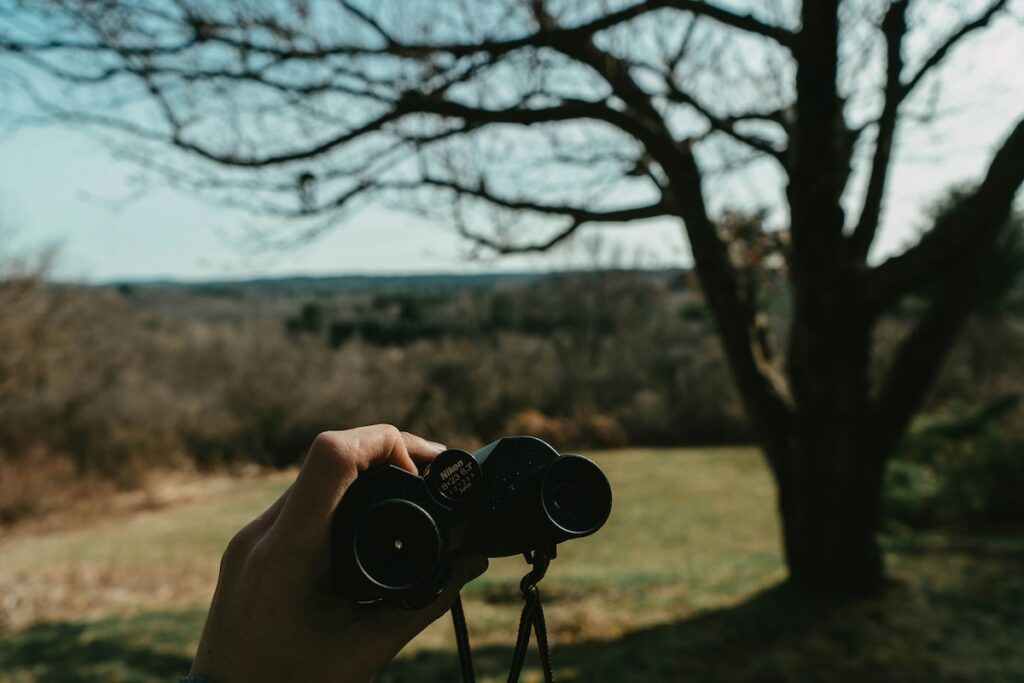 jack-schwartz-llf-ZLTC49M-unsplash | I Heart RVing Close-up of a hand holding binoculars, aimed at a distant rural landscape with leafless trees in the background.