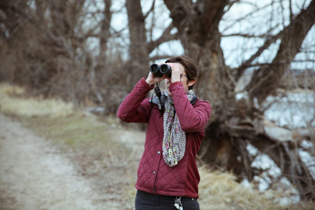 jordan-spalding-Rlpnviei2nc-unsplash | I Heart RVing Woman in a burgundy quilted jacket uses binoculars outdoors among bare trees in a wintry setting, scarf wrapped around her neck.