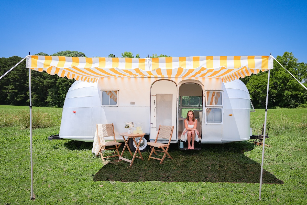 Silver vintage trailer with a yellow striped canopy, a small table and chairs, and a woman sitting on the steps after a full RV renovation.
