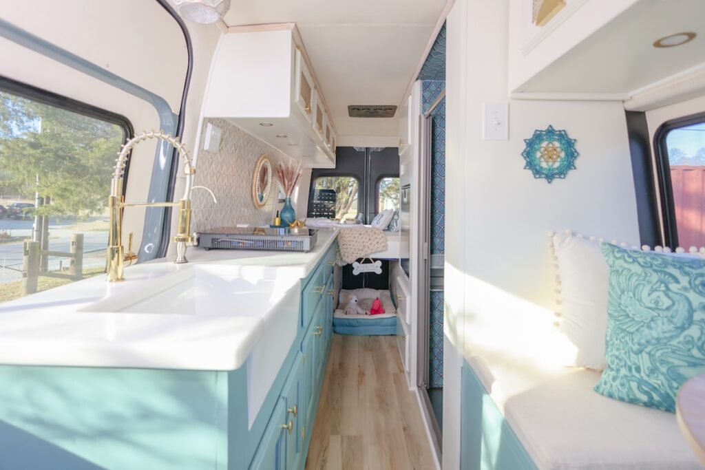 Rise2ShineAthena | I Heart RVing Bright campervan kitchen with teal cabinets, white countertop, and a gold faucet; a seating nook with turquoise pillows visible at right.