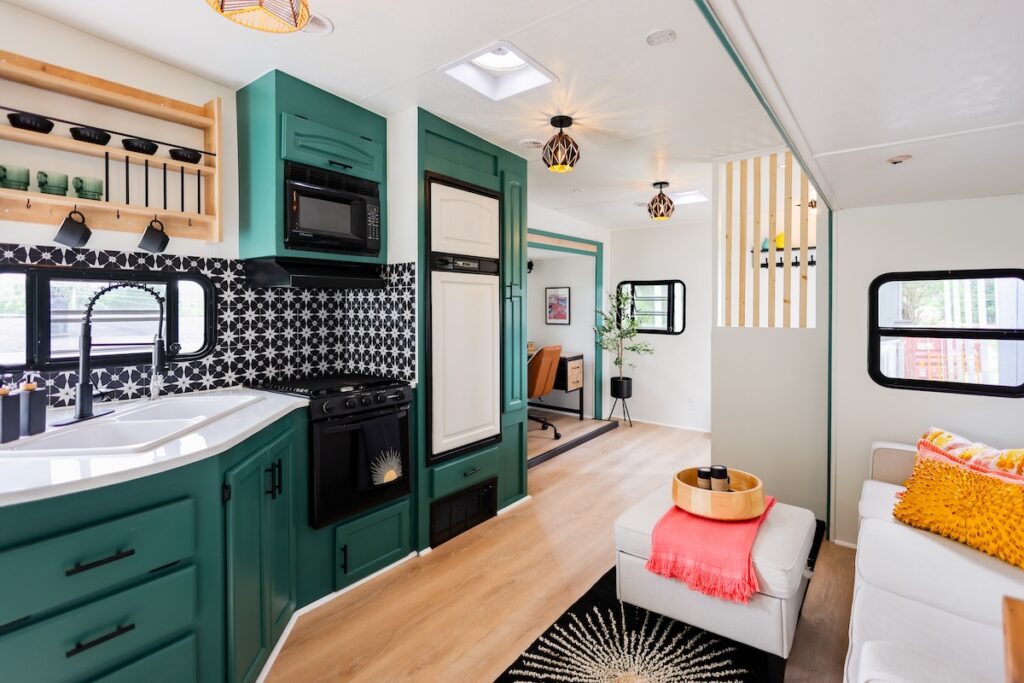 Rise2ShineCricket | I Heart RVing Teal kitchen inside a tiny house with a black-and-white starburst backsplash, white sink, and built-in appliances; open living area with a white sofa and yellow accent pillow visible through a partial divider.