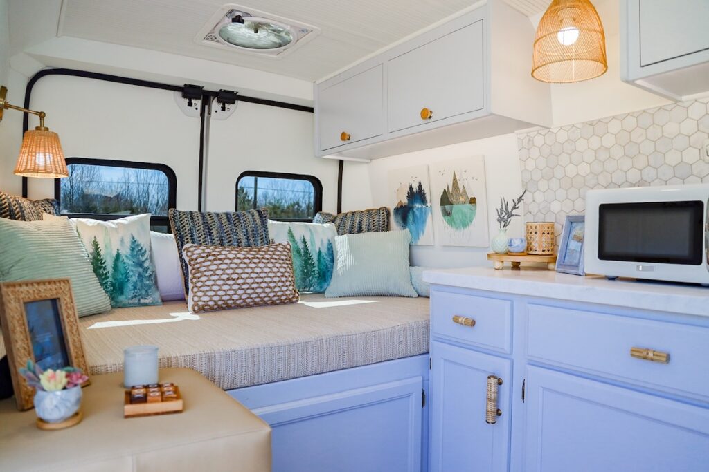 Rise2ShineJuneau | I Heart RVing Cozy camper van living area with a cushioned bench, teal and blue throw pillows, and large windows showing trees outside; light-blue cabinets and a hexagonal backsplash behind a microwave on the counter; wicker pendant lights add warm lighting.