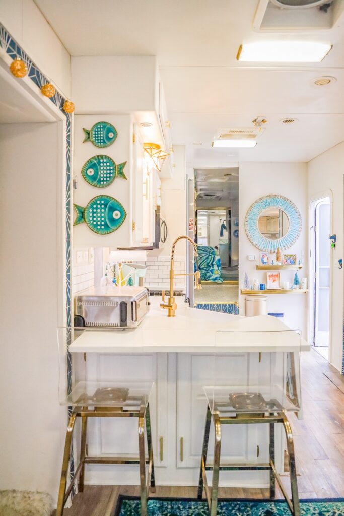 Rise2ShineWander Woman | I Heart RVing Bright coastal kitchen with turquoise fish wall art, white cabinets, and a gold faucet at a breakfast bar.