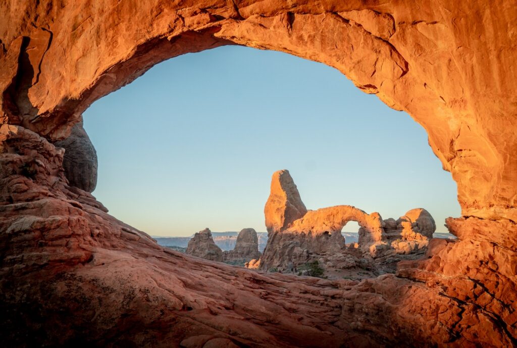 intricate-explorer-EPyKdwWz67k-unsplash | I Heart RVing Sunlit red sandstone arch framing a desert landscape with distant rock formations under a clear blue sky.