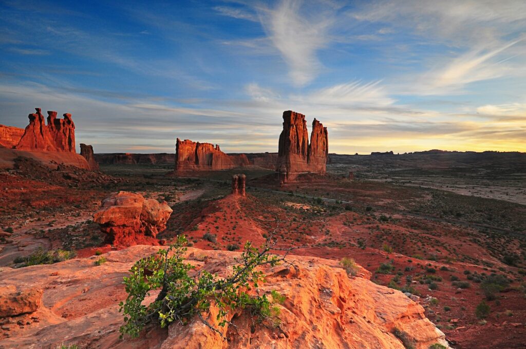 noaa-iDyXVQ1FTgs-unsplash | I Heart RVing Red desert landscape at sunset with tall sandstone formations and a blue sky, foreground showing a green shrub on orange rock.