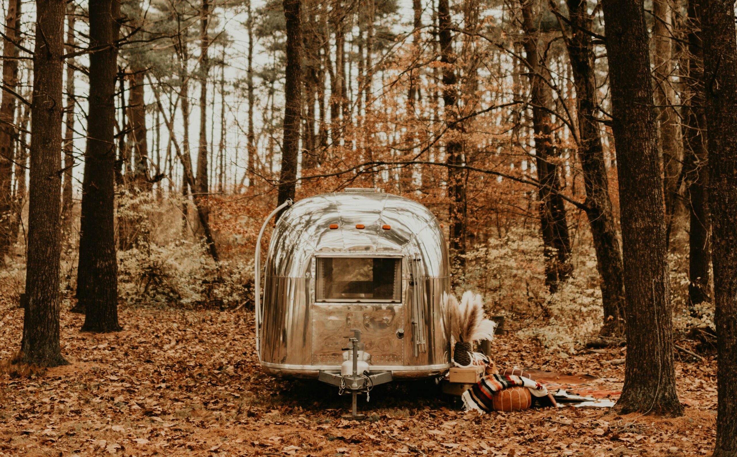 Polished silver vintage travel trailer parked in a forest with autumn leaves, pumpkins, and a striped blanket nearby.